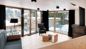 Ein modernes Wohnzimmer mit einer Couch, einem Sessel und einem kleinen Tisch. Große Fenster bieten Blick auf eine verschneite Landschaft und ein Gebäude.