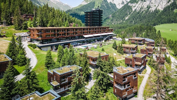 Ein moderner Hotelkomplex mit mehreren Gebäuden, darunter ein markanter Turm, inmitten einer bergigen Landschaft mit üppiger Vegetation und blauem Himmel.