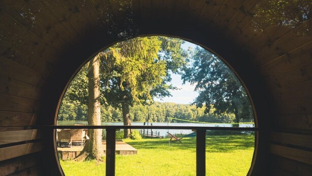 Een rond houten raam met uitzicht op een grasveld, een steiger en een meer.
