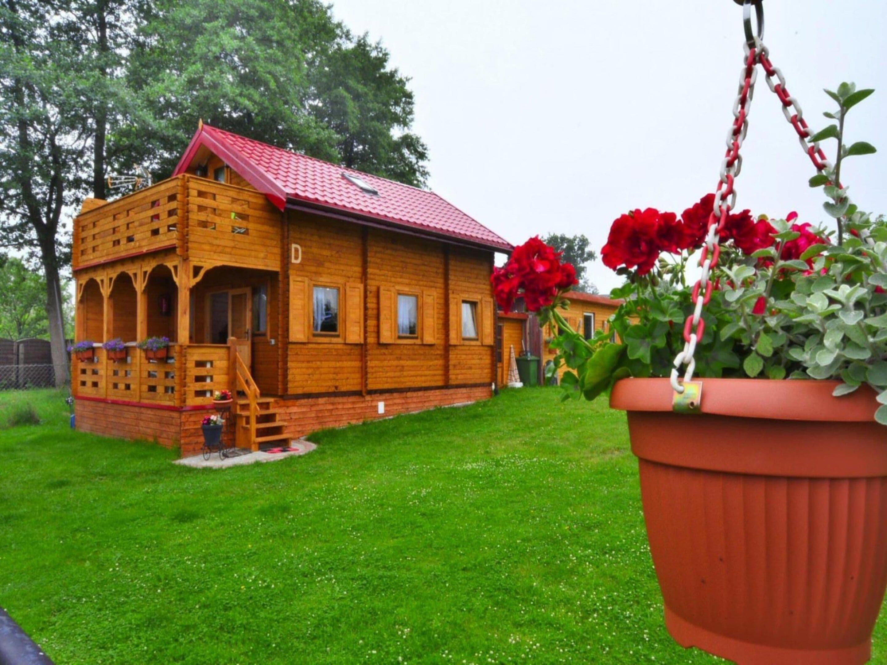 A wooden cabin with a red roof, a porch with a staircase, and a hanging flower pot.