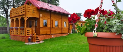 A wooden cabin with a red roof, a porch with a staircase, and a hanging flower pot.