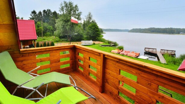 A wooden deck with green chairs, a small building with a red roof, and a view of a lake. - Wooden House by Ostrowieckie Lake With Beach (Malechowo)