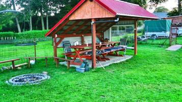A wooden pavilion with a red roof, a fire pit, and a picnic table with chairs.