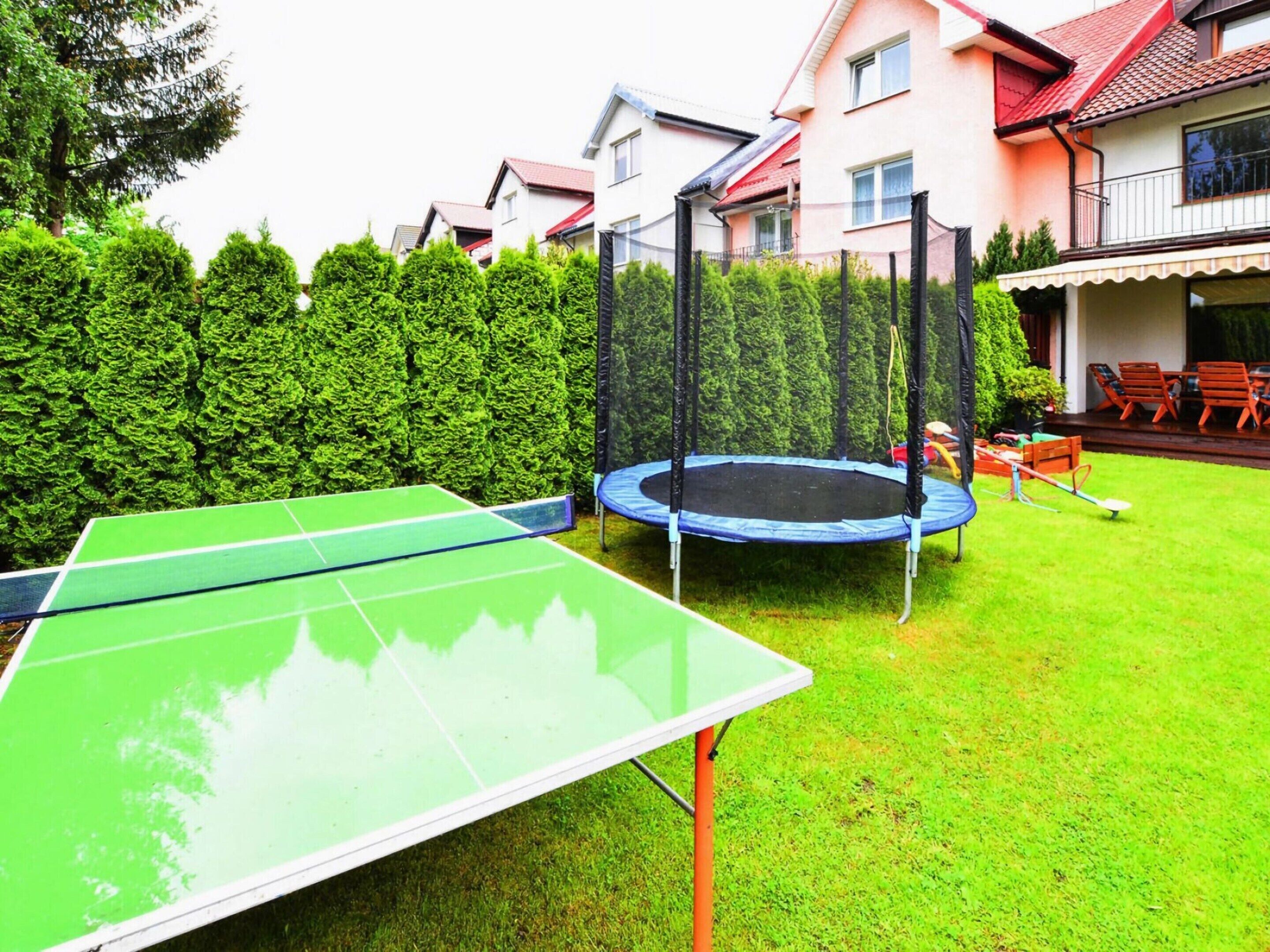 Ein grüner Tischtennis-Tisch und ein Trampolin in einem Garten mit einer Hecke und einem Haus im Hintergrund.