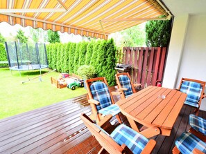 A wooden deck with a table and chairs, a trampoline, and a garden area with a fence and greenery. - Comfortable Holiday Villa, Private Garden (Kolobrzeg)