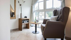 Un salon moderne doté d’une grande fenêtre, d’un fauteuil marron, d’une petite table ronde et d’une étagère en bois sur laquelle se trouvent un appareil photo et des haut-parleurs.