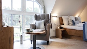 Une chambre moderne avec une grande fenêtre, un fauteuil gris, une table ronde et un canapé agrémenté de coussins à motifs.
