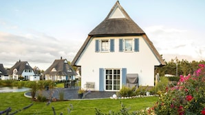 Une maison blanche avec un toit à forte pente et des volets bleus, entourée d’un jardin bien entretenu où poussent diverses plantes et fleurs.