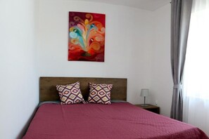 Ein Schlafzimmer mit einem Holzkopfteil, einer roten Bettdecke, gemusterten Kissen, einem Nachttisch mit Lampe und einem farbenfrohen abstrakten Gemälde an der Wand.