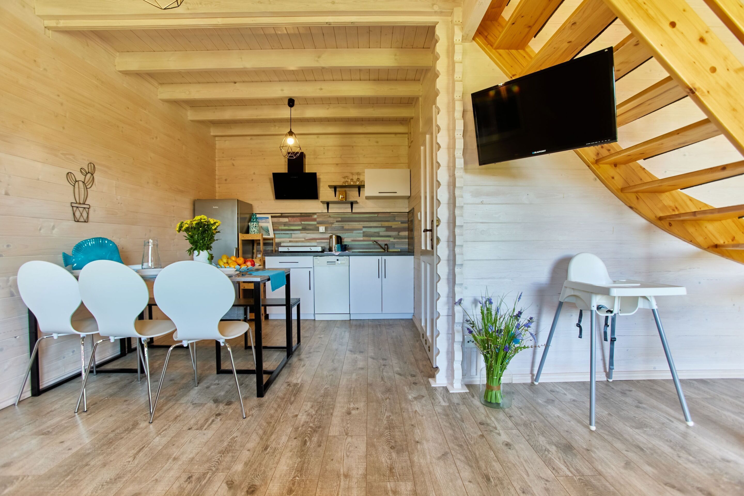A modern kitchen with wooden beams, a dining area with white chairs, and a TV mounted on a wooden beam.