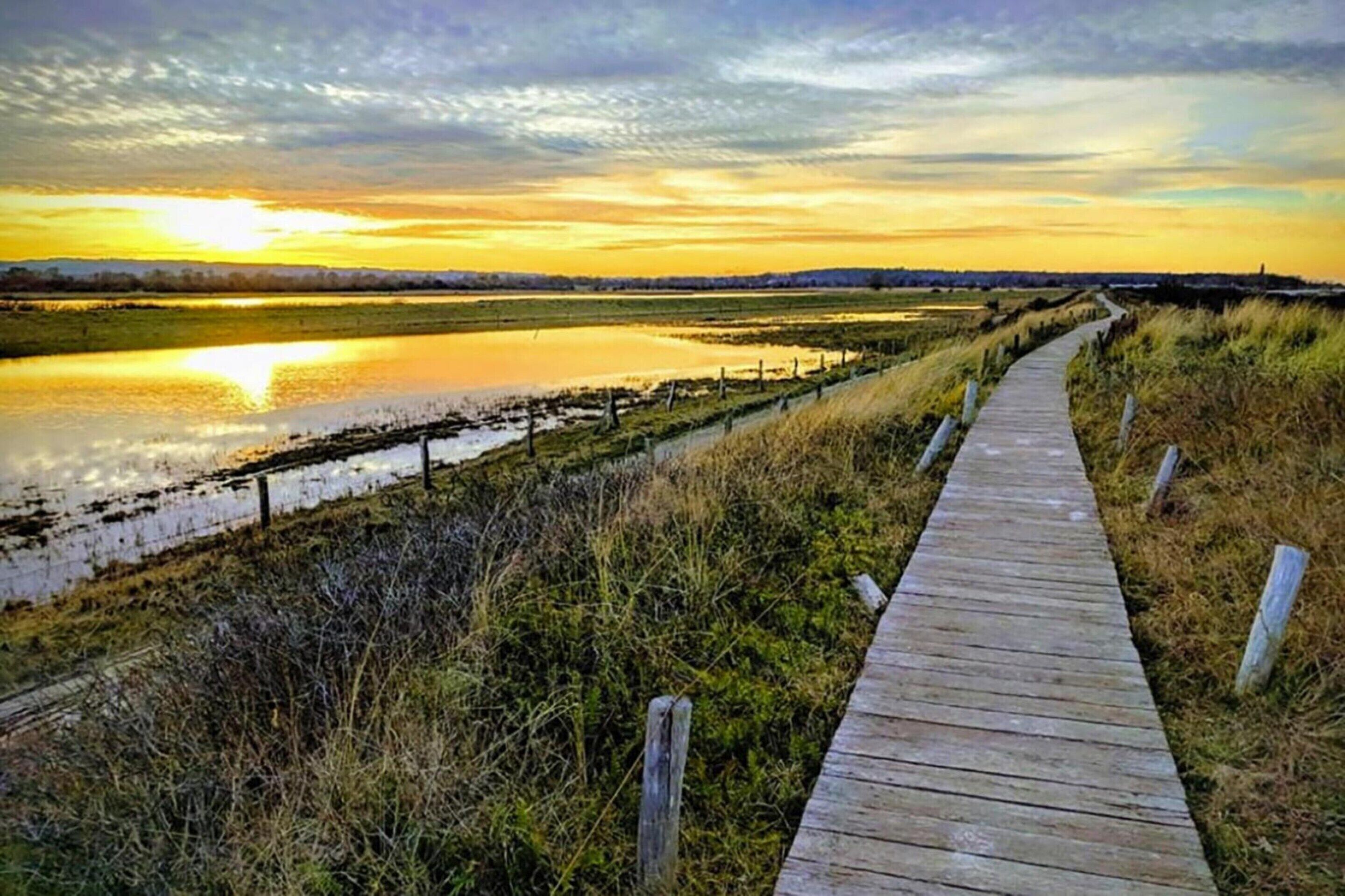 A wooden boardwalk along a water body with grassy banks and a cloudy sky.