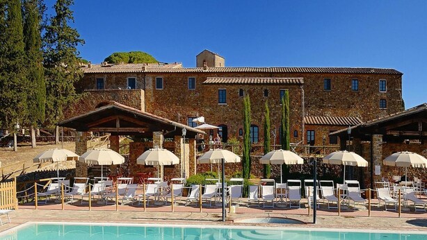 A pool area with lounge chairs and umbrellas, surrounded by stone buildings with multiple windows. - Apartment in Campiglia Marittima (Campiglia Marittima)
