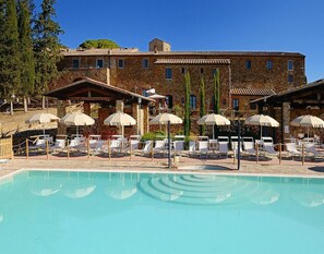 A pool area with lounge chairs and umbrellas, surrounded by stone buildings with multiple windows. - Apartment in Campiglia Marittima (Campiglia Marittima)