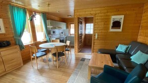 Una cabaña acogedora con interior de madera, un comedor con sillas y una mesa, un sofá, y una cocina visible a través de una puerta abierta.