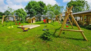 A playground with swings and a sandbox in a grassy area with wooden structures and houses in the background. - Apartment in a Family Holiday Resort (Postomino)