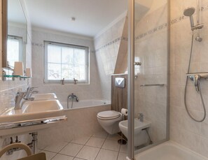 A modern bathroom with a bathtub, a shower, and a toilet. There is a window with blinds and a mirror above the sink. - Ferienwohnung in St. Peter-ording (St. Peter-Ording)