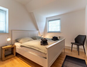 A bedroom with a bed, bedside lamp, chair, and two windows. - Ferienwohnung in St. Peter-ording (St. Peter-Ording)