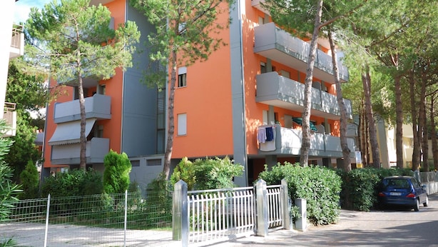 Un edificio residencial de varios pisos con balcones, rodeado de árboles y una valla.