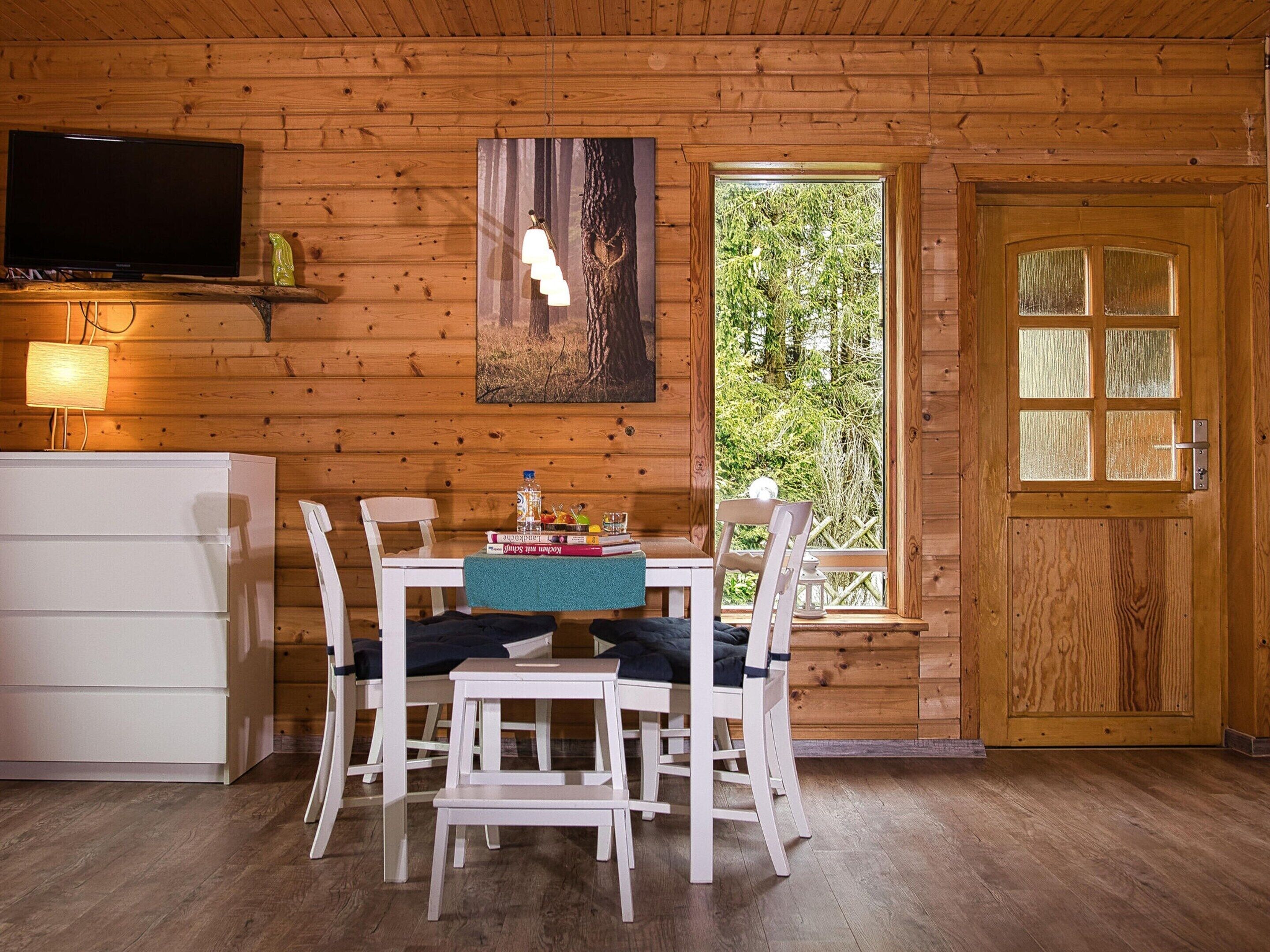 Interior de una cabaña de madera con una mesa de comedor, sillas, un televisor y una puerta con paneles de vidrio.