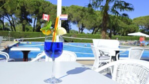Un vaso transparente con una bebida azul, hojas de menta y una bandera, sobre una mesa blanca cerca de una piscina con sillas y palmeras al fondo.