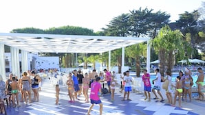 Zona de piscina con piso a cuadros, sombrillas y gente en traje de baño.
