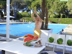 A poolside area with a table displaying watermelon slices and a person raising their hand.