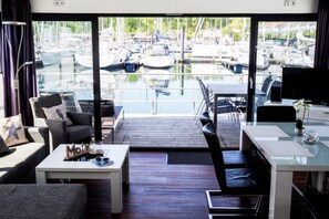 Ein modernes Zimmer mit großem Panoramafenster mit Blick auf eine Marina. Es gibt ein Sofa, einen Esstisch und einen Fernseher.