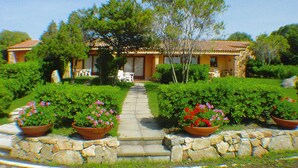 A house with a stone pathway leading to a door, surrounded by well-maintained hedges and flowering plants. - Apartment in Porto Pollo With Garden (Palau)