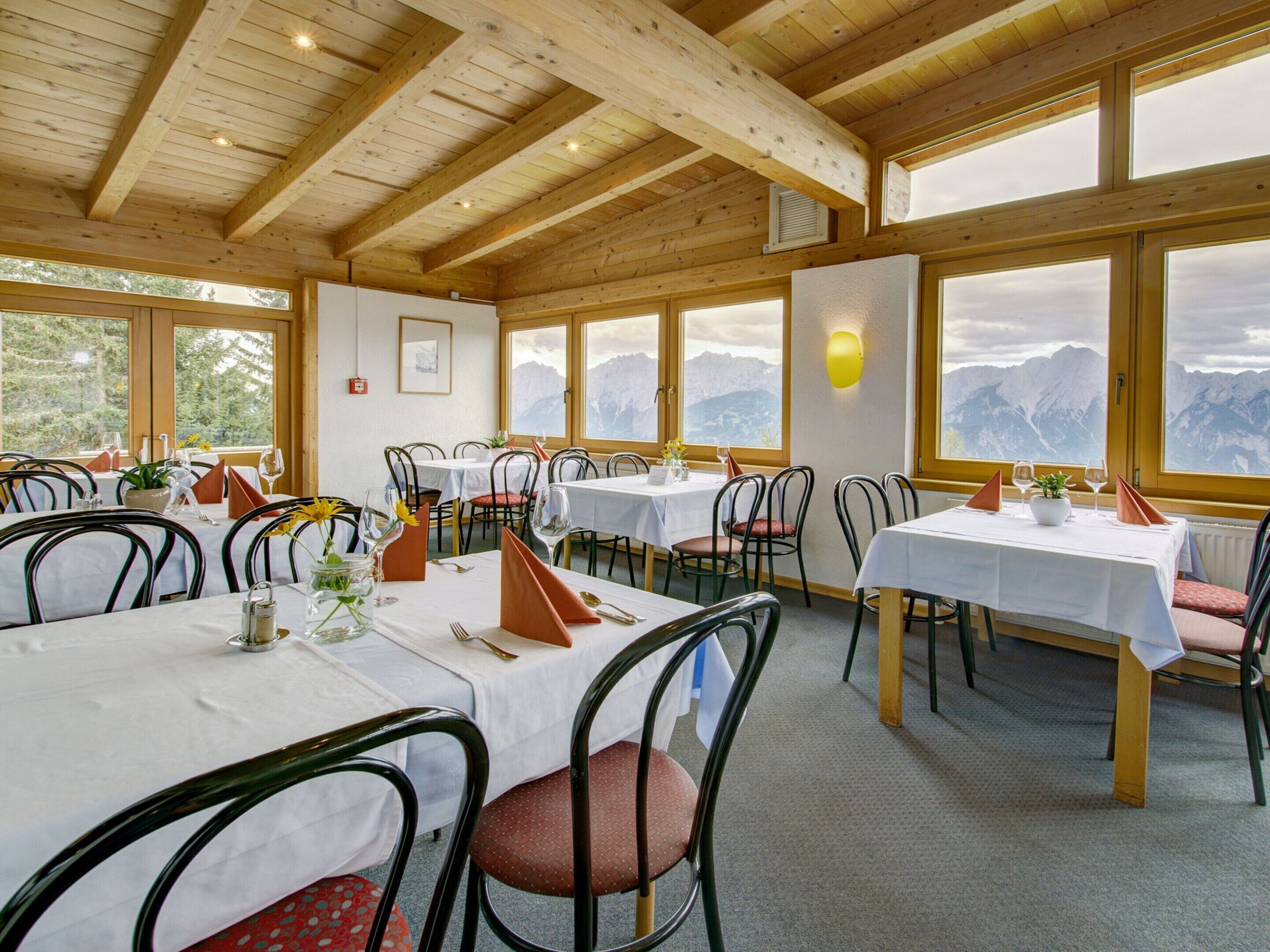 En restaurang med synliga träbjälkar, vita bord och stolar. Den har stora fönster med utsikt över bergen.