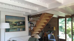 Ein gemütliches Wohnzimmer mit einem weißen Sofa, einem Gemälde, einer Holztreppe und Blick ins Grüne durch große Fenster.