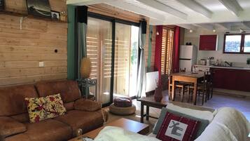 A cozy living room with a brown leather sofa, a green armchair, and a wooden coffee table. There is a kitchen area with a dining table and red cabinets.