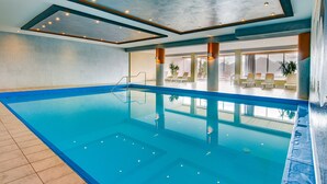 A large indoor swimming pool with clear blue water, a tiled floor, and a ceiling with recessed lighting. There are lounge chairs and potted plants around the pool area. - Apartment in Lienz With a Balcony (Thurn)