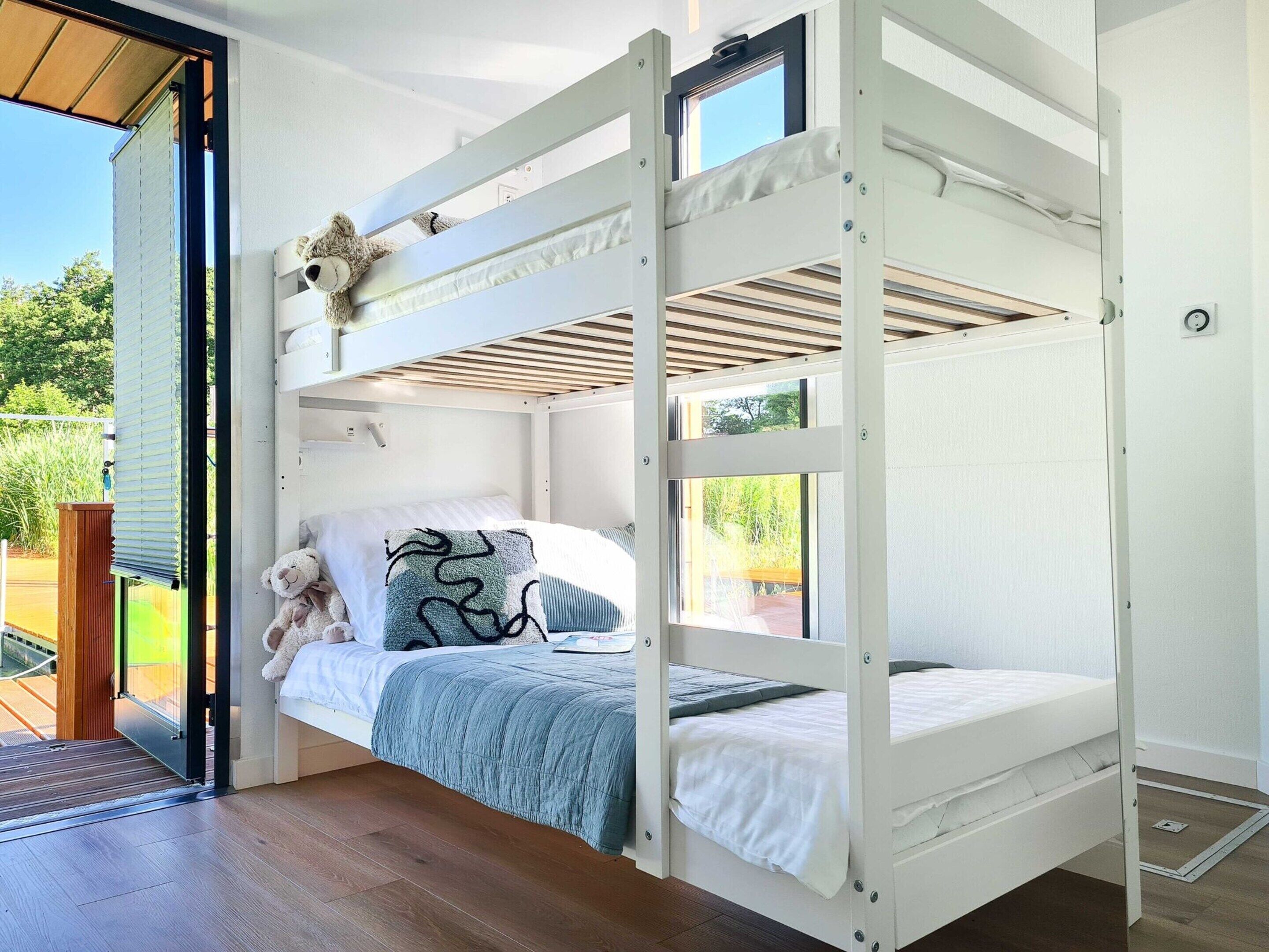 Uma cama beliche com um brinquedo de pelúcia e almofadas estampadas, uma janela com vista para o verde e um piso de madeira.