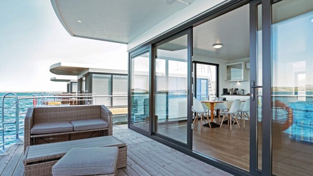 Eine moderne Außenterrasse mit Rattan-Sofa und Hocker, einem Essbereich mit Tisch und Stühlen sowie einem gläsernen Anbau mit Meerblick.