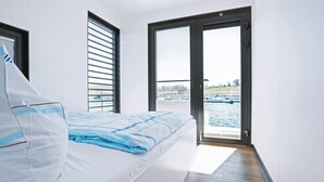 Ein Schlafzimmer mit einem Bett, einem Fenster mit Jalousien und einer Tür, die auf einen Balkon mit Blick auf Wasser und Boote führt.