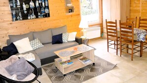 Une pièce chaleureuse aménagée dans un chalet en rondins, avec un canapé, une table en bois, une chaise, une lampe et une fenêtre donnant sur un paysage verdoyant.