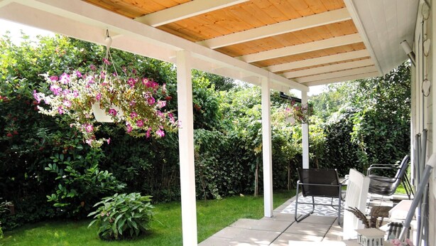 Un patio couvert avec un plafond en bois, une composition florale suspendue et un espace détente avec des chaises et une table.