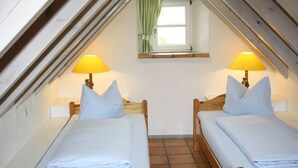 Ein kleines, gemütliches Schlafzimmer im Dachgeschoss mit zwei Einzelbetten, jeweils mit einem Holzkopfstück und einem Nachttisch. Ein Fenster mit Vorhang und eine Lampe befinden sich auf jeder Seite des Raumes.