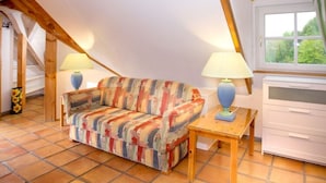Ein gemütliches Dachzimmer mit einem farbenfrohen Sofa, zwei Lampen, einem kleinen Tisch und einem Fenster mit Blick auf Grünflächen.