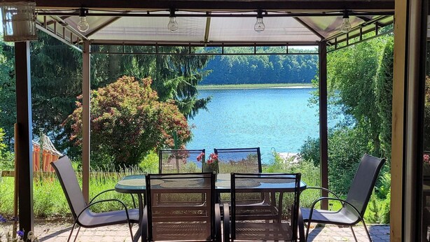 A covered patio with a table and chairs, overlooking a lake and surrounded by greenery. - Holiday Home Right on the Lake, Florczaki (Lukta)