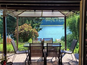 A covered patio with a table and chairs, overlooking a lake and surrounded by greenery. - Holiday Home Right on the Lake, Florczaki (Lukta)