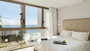 Une chambre d’hôtel dotée d’une grande fenêtre donnant sur la mer, d’un lit recouvert d’une couette blanche et agrémenté d’oreillers à motifs, et d’un plateau de petit-déjeuner posé sur le lit.