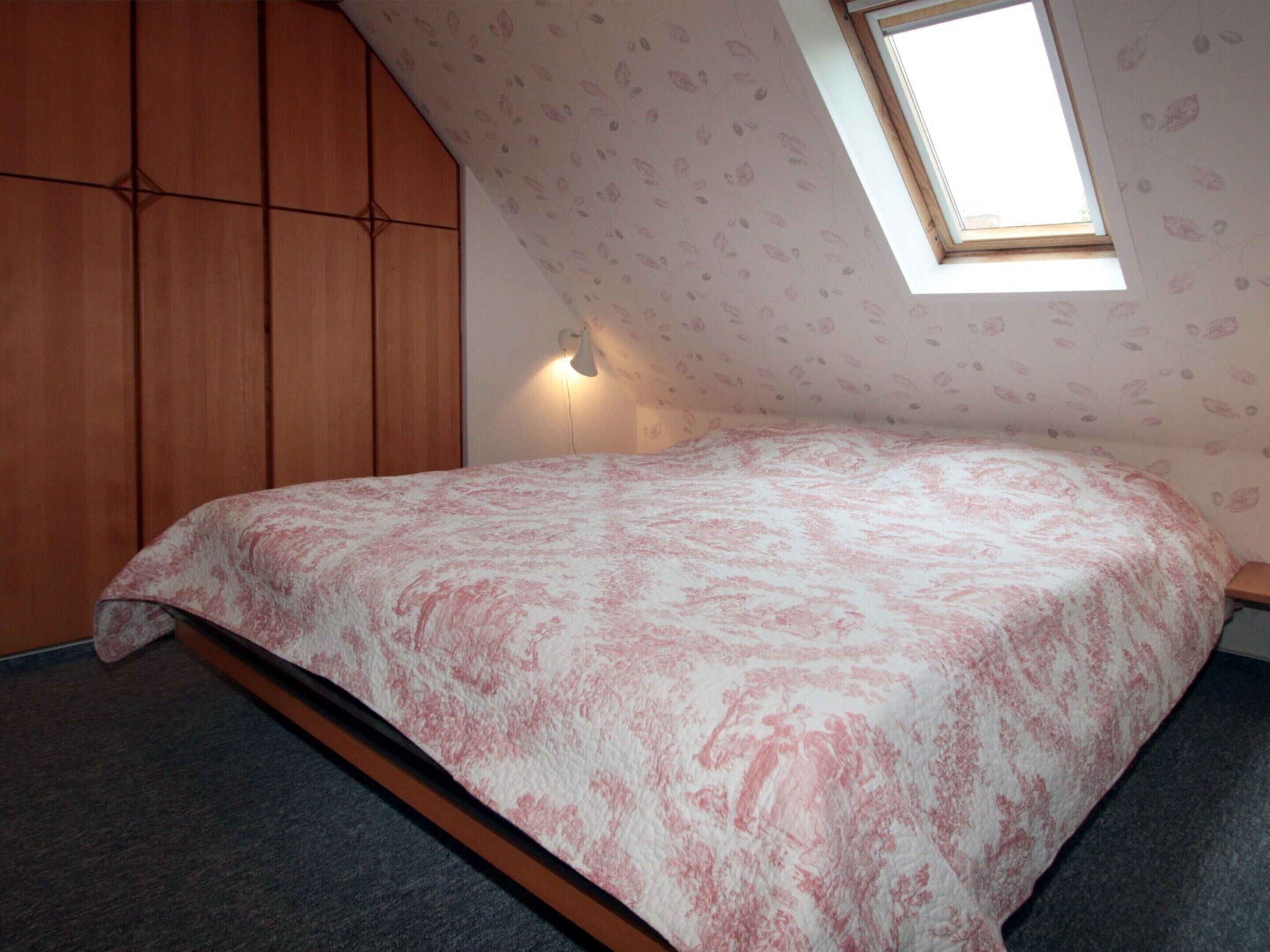 Ein Schlafzimmer mit schrägen Wänden, einem Bett mit Blumenmuster, einem Holzschrank und einem kleinen Fenster mit Aussicht.