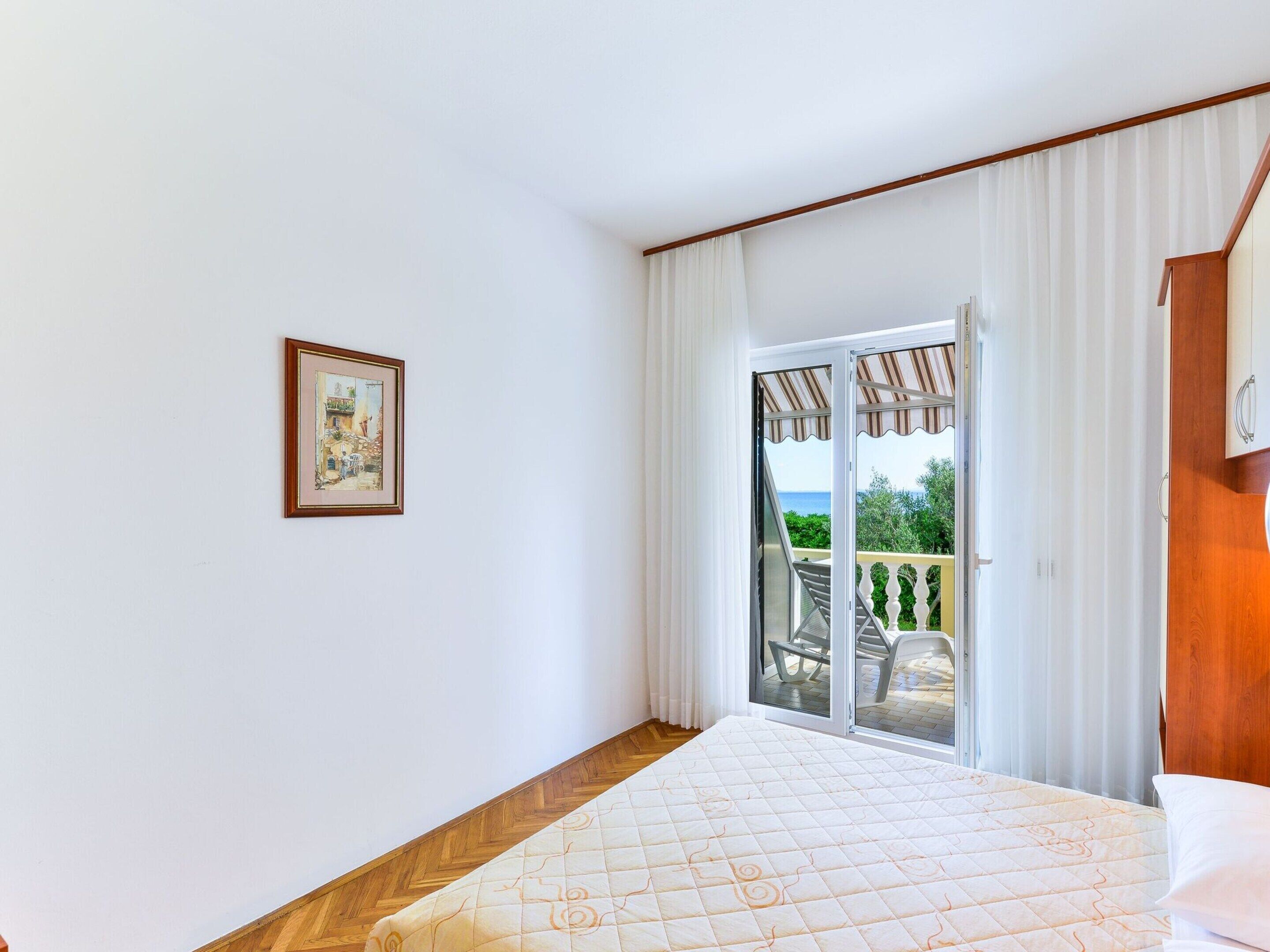 A bedroom with a bed, a framed picture on the wall, and a sliding glass door leading to a balcony with a view of the sea.