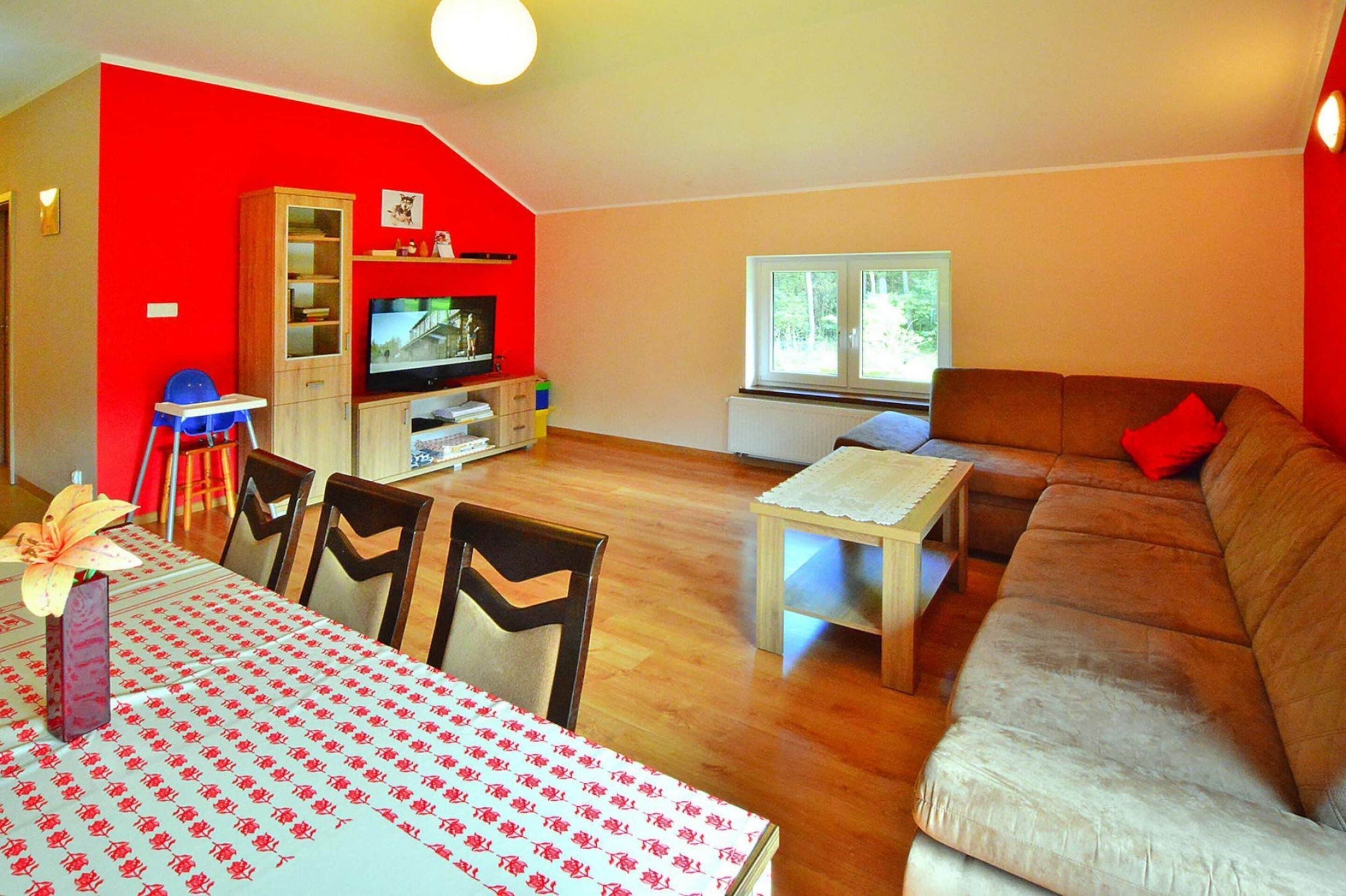 A living room with a red wall, a brown sofa, a wooden dining table with chairs, a television, and a window with a view of trees.