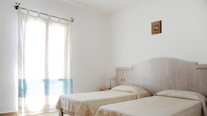 Une chambre avec deux lits, une fenêtre avec des rideaux légers, une table de chevet avec une lampe et un tableau encadré au mur.