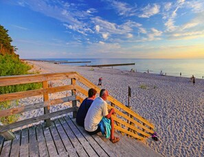 A wooden boardwalk leading to a sandy beach with a clear view of the ocean and a wooden pier extending into the water. - Holiday Home in Sianozety Near the Beach (Ustronie Morskie)