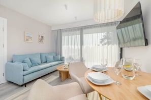 A modern living room with a sofa, a dining table set with plates and glasses, and a flat-screen TV mounted on the wall.