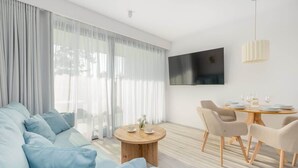A modern living room with a sofa, a round wooden coffee table, a dining table with chairs, and a flat-screen TV mounted on the wall.