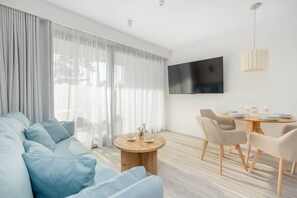 A modern living room with a sofa, a round wooden coffee table, a dining table with chairs, and a flat-screen TV mounted on the wall.
